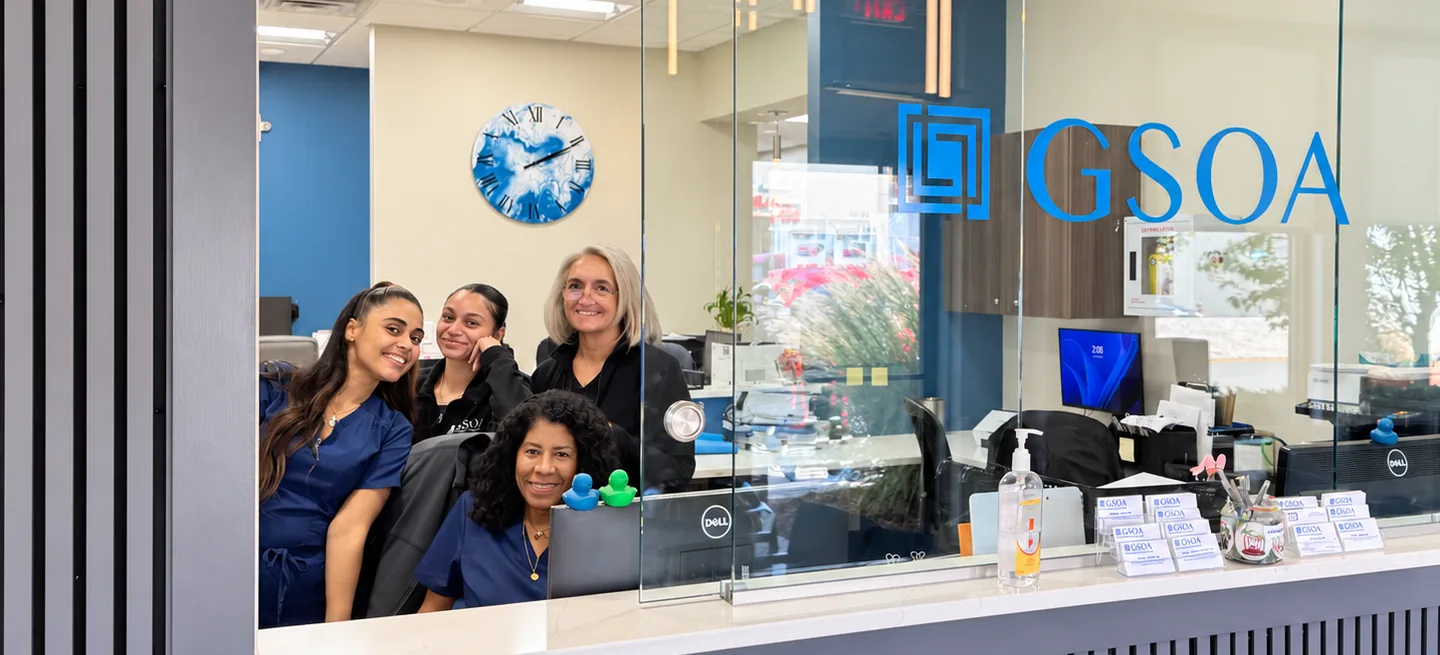 Smiling members of the Garden State Orthopaedic Associates front desk team behind the reception window, with the GSOA logo on the glass